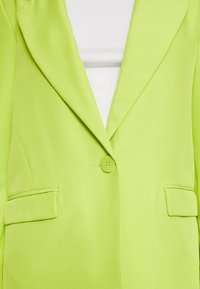 Limonkowo-zielona marynarka o gładkiej fakturze, z wyciętymi klapami i zapięciem na jeden guzik. Posiada dwie przednie kieszenie.