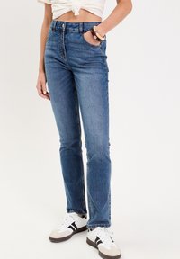 Blå denim jeans med høj talje, slim fit, subtil falming og fem lommer, parret med hvide sneakers med sorte striber.