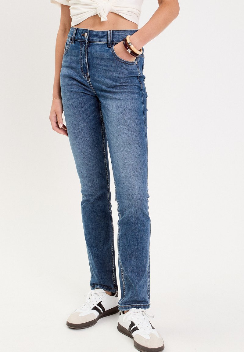 Blå denim jeans med høj talje, slim fit, subtil falming og fem lommer, parret med hvide sneakers med sorte striber.