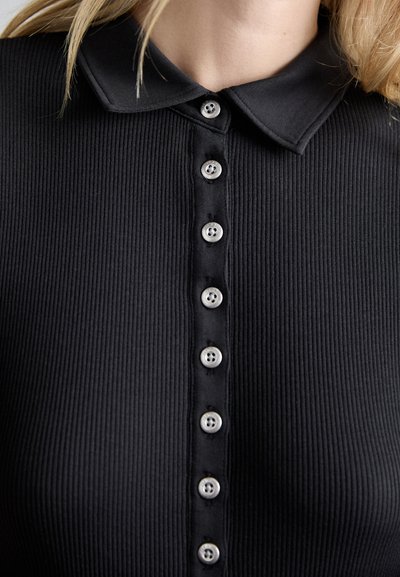 Top nero a coste con colletto e una fila di otto bottoni argentati sul davanti. Il tessuto ha un aspetto testurizzato.