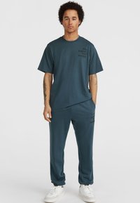 Teal bombažna mešanica, ki vključuje t-shirt sproščenega kroja s sprednjim potiskom in usklajene trenirke, kombinirane z belimi supergami.