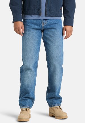 Man die een blauwe jeans, beige suède laarzen en een donkerblauwe jas draagt, staande tegen een witte achtergrond.