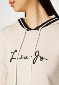 Sweat à capuche beige avec cordon de serrage rayé noir et blanc et logo "Liu Jo" brodé en noir. Tissu doux avec une l égère texture brillante.