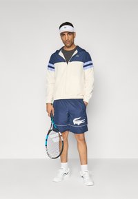 Marineblauwe en crèmekleurige windbreaker met gestreepte accenten, gecombineerd met marineblauwe shorts met een wit krokodillenlogo. Houdt een tennisracket vast.