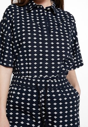 Femme portant une chemise bleu marine à manches courtes avec boutons et un pantalon assorti à motif géométrique blanc, taille élastique avec cordon.