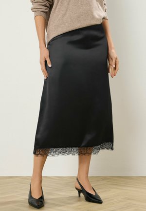Kobieta w czarnej, satynowej spódnicy midi z koronkowym wykończeniem, beżowym swetrze oraz czarnych sandałkach na niskim obcasie, stojąca na drewnianej podłodze.