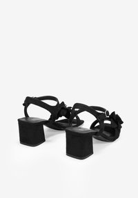 Sandalias de tacón de ante negro con un chunky block heel, parte trasera abierta y adornos florales decorativos en la correa. Suela plana y hebilla ajustable.