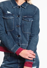 Farkkutakki, jossa on kaksi etutaskua, tummansininen väri ja ribbireunat tummanpunaisena. Sisältää pientä kulumista ja napit keskellä.