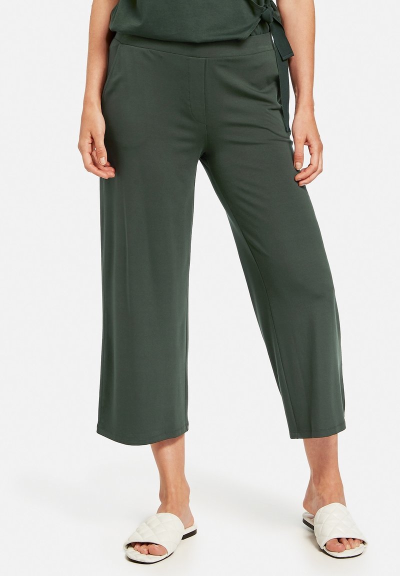 Pantalon léger, vert foncé, à jambes larges, avec une taille élastique et des poches latérales, associé à des sandales blanches matelassées à enfiler.