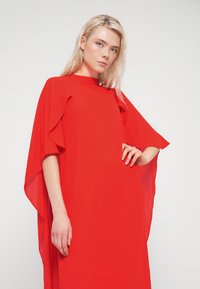 Κόκκινο, ρέον φόρεμα από ελαφρύ ύφασμα με σιλουέτα Α-γραμμή, διαθέτον φαρδιά μανίκια και ψηλό λαιμό για μινιμαλιστικό σχεδιασμό.