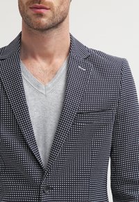 Mężczyzna w granatowej marynarce w wzory nałożonej na jasnoszary sweter typu V-neck, skrócony w okolicy szyi i klatki piersiowej.
