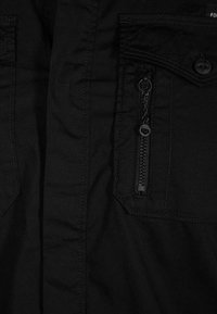 Černá košile z látky s kapsou na zip, dvěma náprsními kapsami s klopami a prošívacími detaily. Hladký povrch a pohodlný střih.