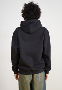 Μαύρη φούτερ με κουκούλα και άνετη εφαρμογή, με μακριά μανίκια και ριμπ μπάσκα. Συνδυασμένο με ανοιχτό μπλε παντελόνι σε φαρδιά γραμμή.