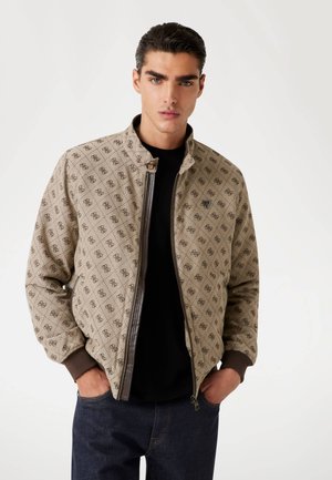 Chaqueta acolchada beige con acabados marrones, que presenta un cuello alto, cierre de cremallera y un patrón geométrico en toda su superficie. Llevada sobre una camisa negra.