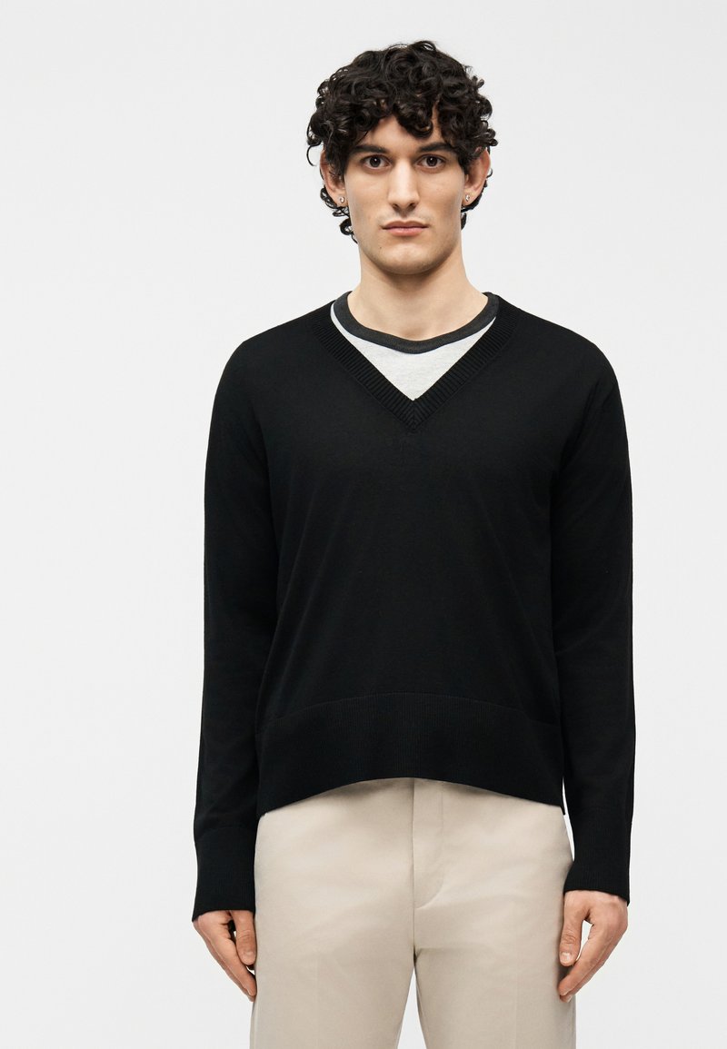 Νεαρός με κατσαρά μαλλιά φορούσε μαύρο πουλόβερ με V λαιμόκοψη πάνω από γκρι πουκάμισο και μπεζ παντελόνι, στέκεται μπροστά σε ένα απλό φωτεινό φόντο.