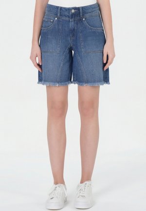Jeansshorts mit vorderen aufgesetzten Taschen und ausgefransten Säumen, getragen von einer Person. Sie tragen weiße Sneaker und ein weißes Oberteil.