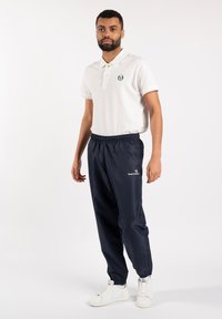 White polo shirt with a collar and a logo on the chest, paired with navy blue track pants and white sneakers.