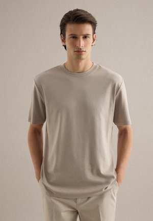 Jonge man met donker haar, gekleed in een beige t-shirt met korte mouwen en lichtgekleurde broek, staand tegen een effen neutrale achtergrond.