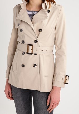 Beige kaksirivinen trenssitakki mustilla napeilla, vyötäröllä vyö ja kalvosinnauhat, päällä raidallinen paita ja harmaat farkut.