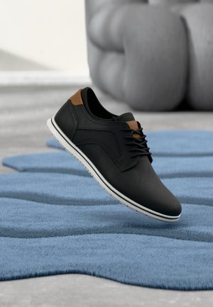 Schwarzer freizeitlicher Lederschuh mit braunen Akzenten, der über einem strukturierten blauen Teppich in einem minimalistischen Innenraum schwebt.