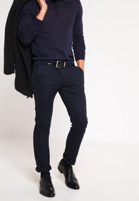 Polo à manches longues bleu marine avec col, pantalon foncé ajusté, et chaussures noires vernies. La tenue comprend un manteau noir posé sur le bras.