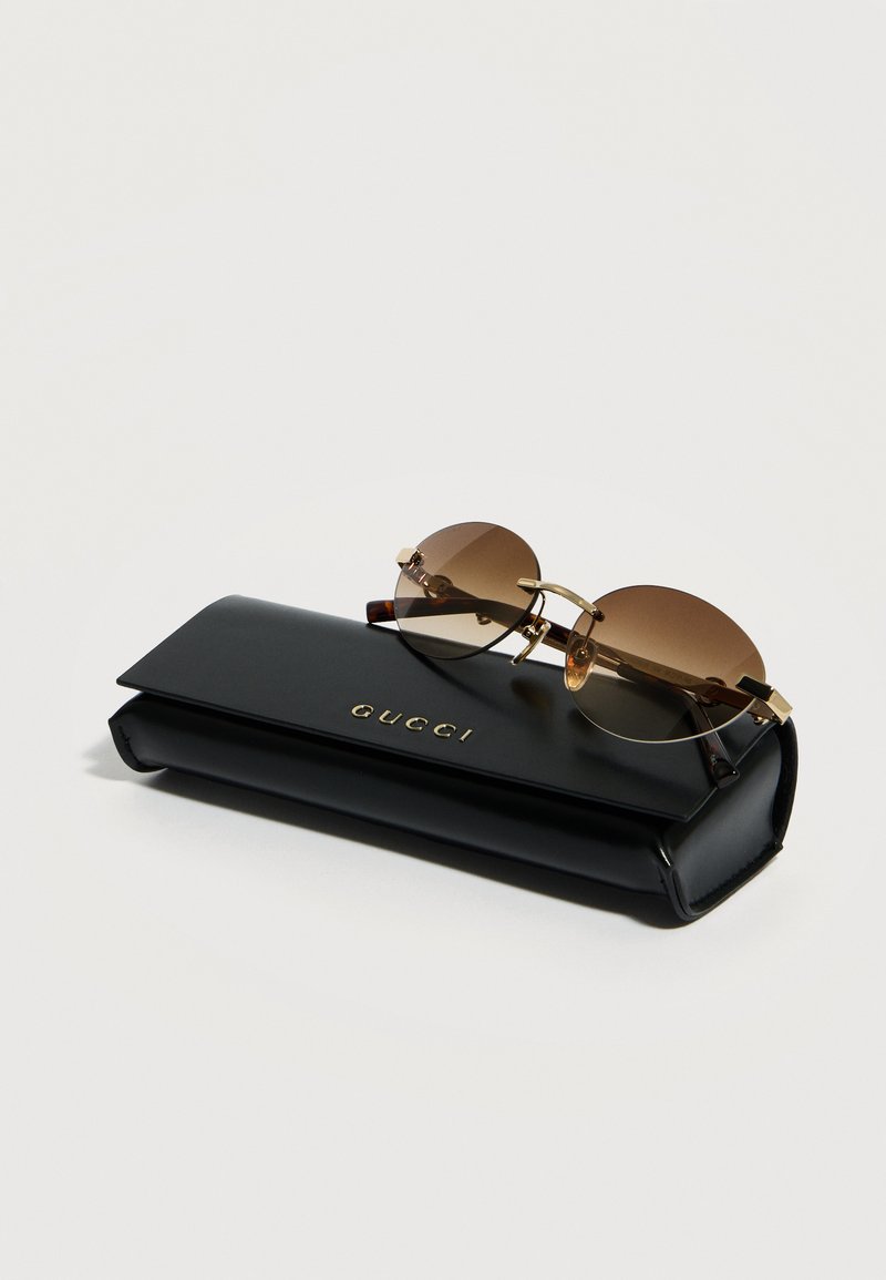 Lunettes de soleil rondes à dégradé marron posées sur un étui à lunettes rectangulaire noir Gucci avec logo doré sur fond blanc.