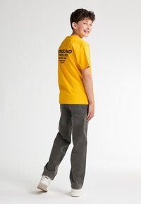 Camiseta de algodón amarilla con texto gráfico negro en la parte de atrás, combinada con pantalones de mezclilla grises y zapatillas blancas, vista en un ángulo.