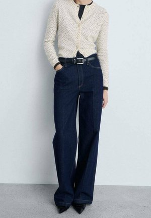 Woman wearing cream polka dot cardigan over black top, high-waisted wide-leg dark blue jeans, black belt, and pointed black shoes.