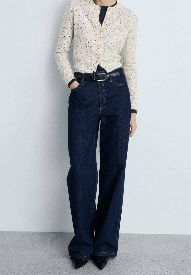 Femme portant un cardigan crème à pois sur un top noir, un jean large taille haute bleu foncé, une ceinture noire et des chaussures noires pointues.