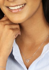 Zilveren ketting met een verstrengeld hartje als hanger, gedragen met een gestreepte lichtblauwe shirt. Soepele ketting en gedetailleerd hartontwerp.