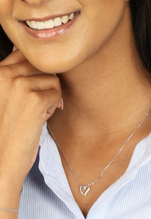 Collier en argent avec pendentif en forme de cœurs entrelacés, porté avec une chemise rayée bleu clair. Chaîne lisse et design de cœur détaillé.