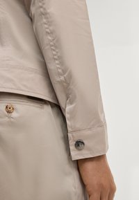 Rękaw beżowej kurtki z mankietem zapinanym na guzik spoczywający blisko kieszeni beżowych spodni z guzikiem, ukazujący szczegółowe przeszycia i teksturę tkaniny.