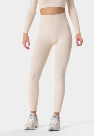 Beige high-waisted legging gemaakt van zacht, rekbaar materiaal met ribbeltextuur en logodetail. Gecombineerd met lichtgekleurde sneakers.