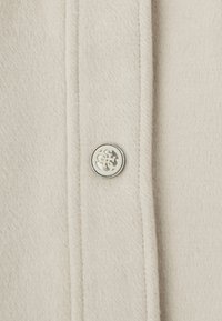 Tejido beige con una textura suave, que presenta un botón plateado con un diseño intrincado, alineado a lo largo de una costura.