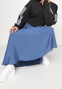 Personne assise portant un haut noir à manches longues avec des rayures blanches, une jupe midi bleue, des baskets blanches montantes et des chaussettes blanches sur un sol gris.