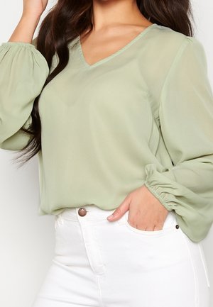 Blouse légère verte claire, transparente, avec un col en V et des manches longues bouffantes. Texture douce, coupe décontractée, assortie à un jean blanc.