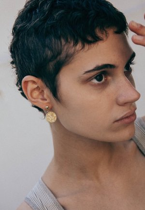 Des boucles d'oreilles en forme de pièce d'or présentent une surface texturée et une petite tige ronde. Le modèle a les cheveux courts et bouclés et porte un débardeur gris.