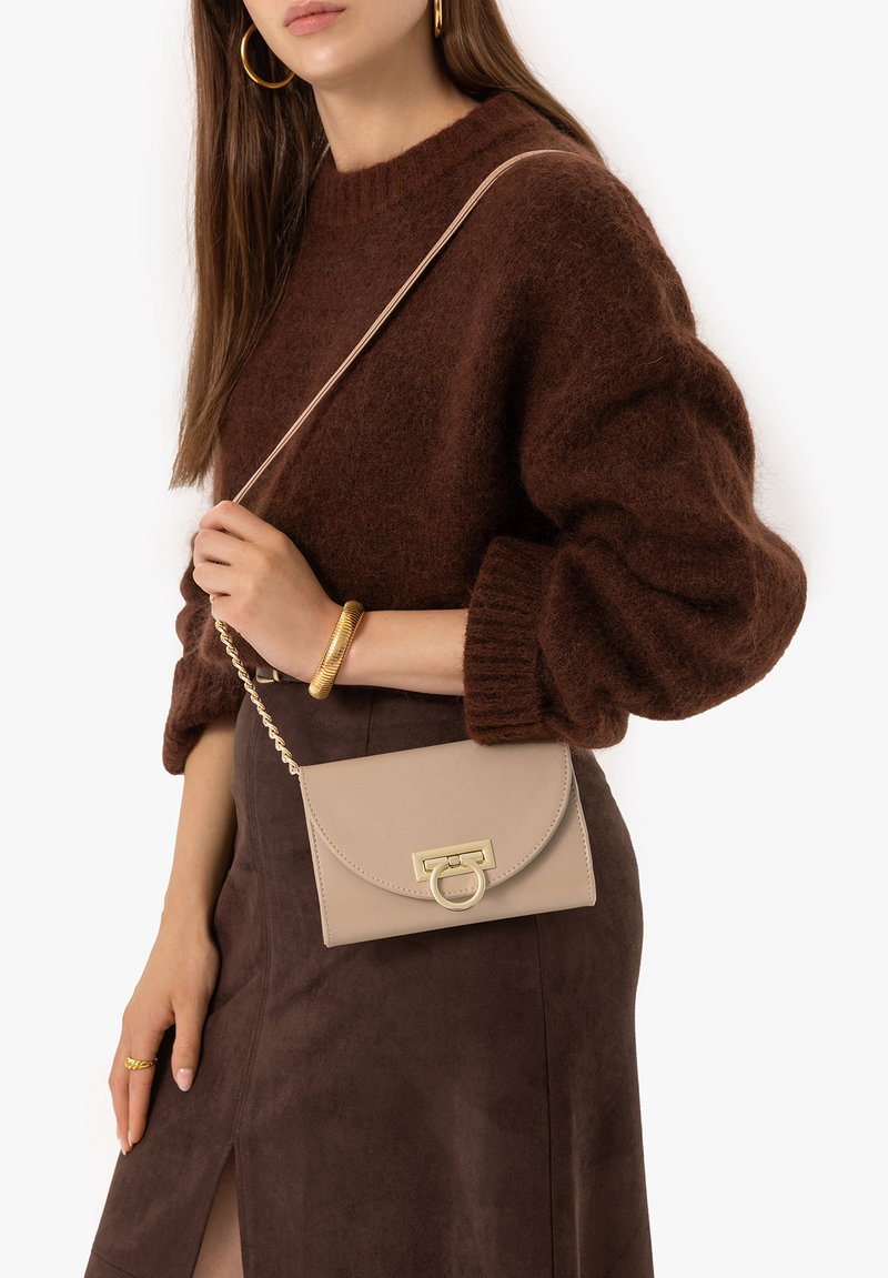 Femme portant un pull marron duveteux et une jupe, tenant un sac bandoulière beige à fermoir doré, accessoirisée de créoles dorées et d'un bracelet.