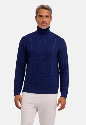 Hombre con cabello gris que lleva un suéter de cuello alto de punto trenzado azul oscuro y pantalones blancos, de pie frente a un fondo liso.
