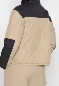 Vadderad jacka med en svart övre del och en beige nedre del. Slät tyg med hög krage och elastisk nederkant.