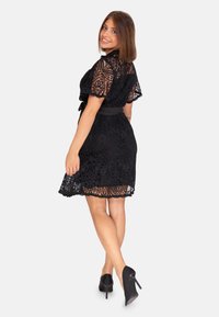 Robe en dentelle noire avec des manches courtes et un ourlet à volants, ornée de motifs floraux complexes et d'une ceinture en tissu à la taille, associée à des talons hauts.