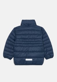Veste matelassée bleu marine avec un col haut, texture en piquage et un détail réfléchissant au dos près de l'ourlet. Fermetures à boutons au niveau du col.