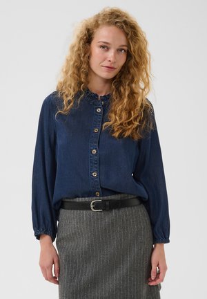 Femme aux cheveux blonds bouclés portant un chemisier bleu foncé boutonné et une jupe grise à fines rayures avec une ceinture noire, debout devant un fond uni.