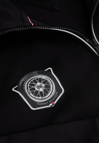 Čierna tkanina so strieborným a čiernym kruhovým emblémom s dizajnom kolesa, logom Cadillac a značkou Tommy Hilfiger blízko zipsu.