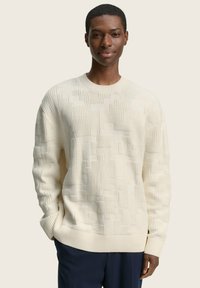 Cremfarbener Strickpullover mit strukturiertem Muster, gerippten Bündchen und Saum. Das Model trägt eine marineblaue Hose. Heller Hintergrund.