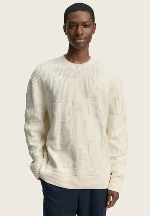 Cream knit sweater with textured pattern, ribbed cuffs, and hem. Model wears navy trousers. Light background.