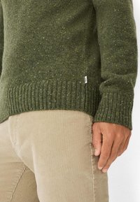 Olivgrüner Strickpullover mit strukturierter Oberfläche und geripptem Saum. Getragen über hellbeigen Cordhosen, mit einer Hand, die seitlich ruht.