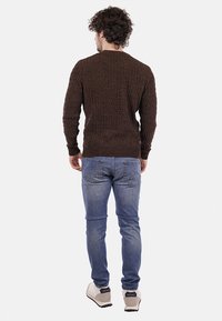 Maglione marrone lavorato a maglia con un motivo testurizzato, maniche lunghe e polsini a coste, abbinato a jeans blu e sneakers di colore chiaro.