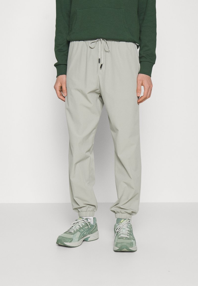 Person med beige bukser med elastisk ærme, grønne sneakers og en mørkegrøn sweatshirt foran en neutral hvid baggrund.