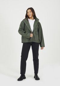 Veste imperméable verte avec capuche, coupe oversize, boutons-pression, deux poches avant, portée par-dessus une chemise blanche et associée à un jean noir et des bottes.